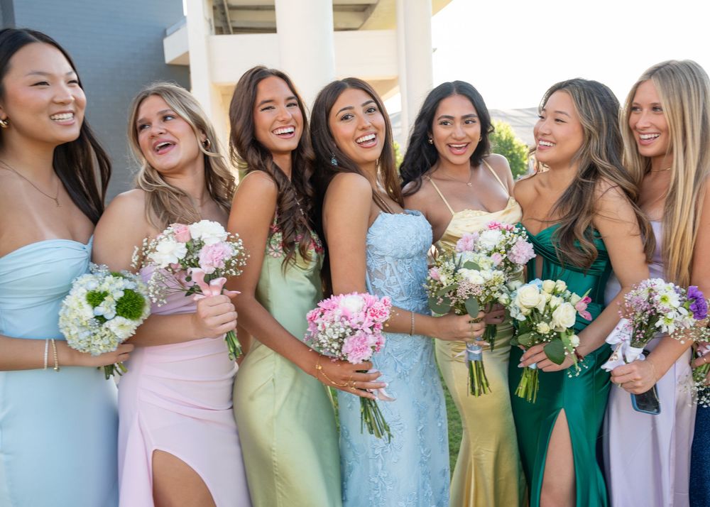 Seven bridesmaids in pastel and colorful dresses laugh together holding beautiful floral bouquets.