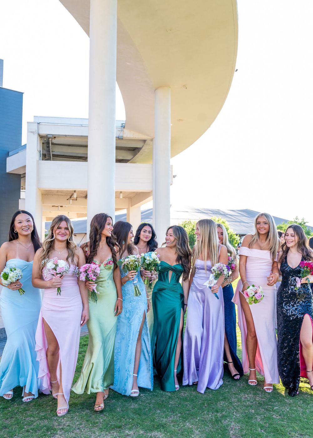 Nine bridesmaids pose in colorful mismatched formal gowns holding bouquets at an outdoor venue.