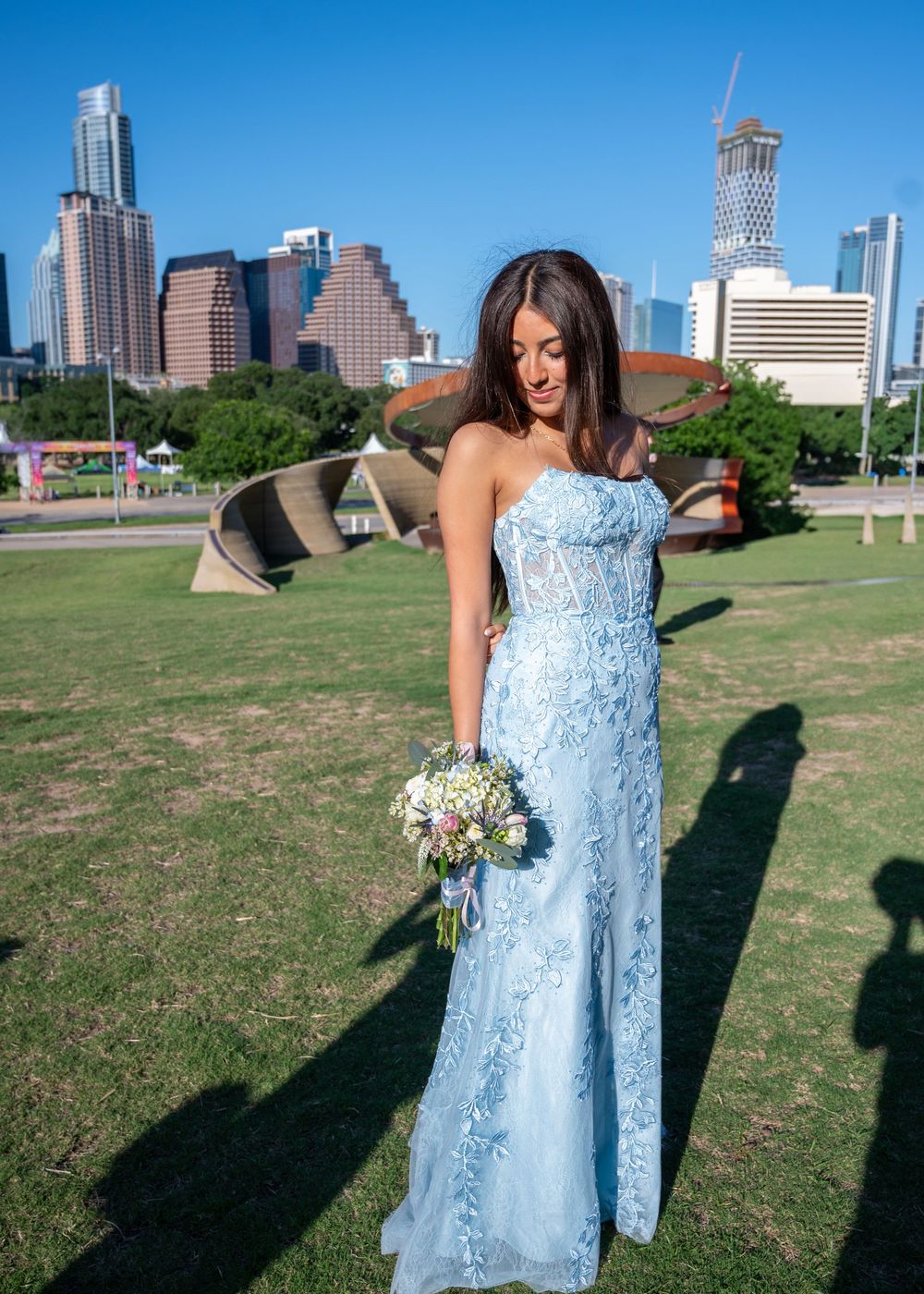 A woman in a strapless light blue lace gown holds a bouquet with an Austin city skyline backdrop.