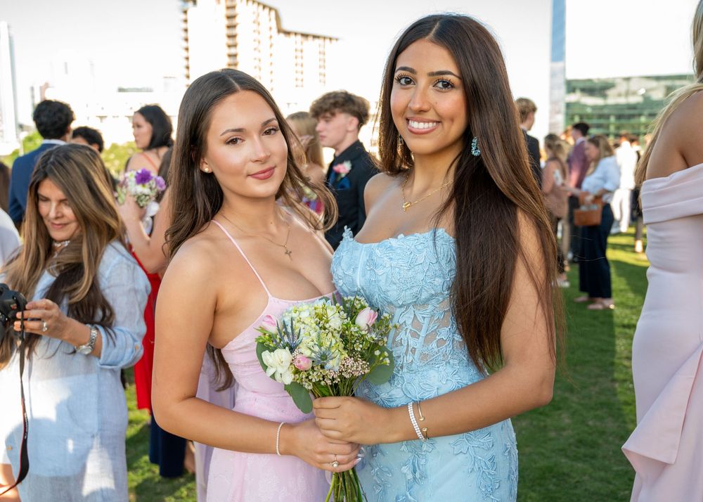 Two young women in pastel prom dresses smile together holding a floral bouquet outdoors.