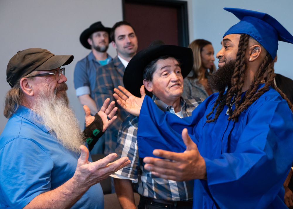 Graduate in blue cap and gown celebrating with guests at an indoor graduation ceremony.