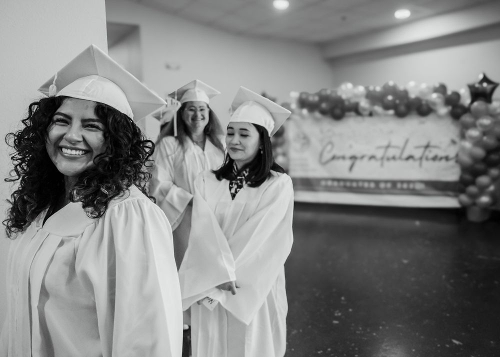 Three women in white graduation caps and gowns smile before a Congratulations banner in black and white.