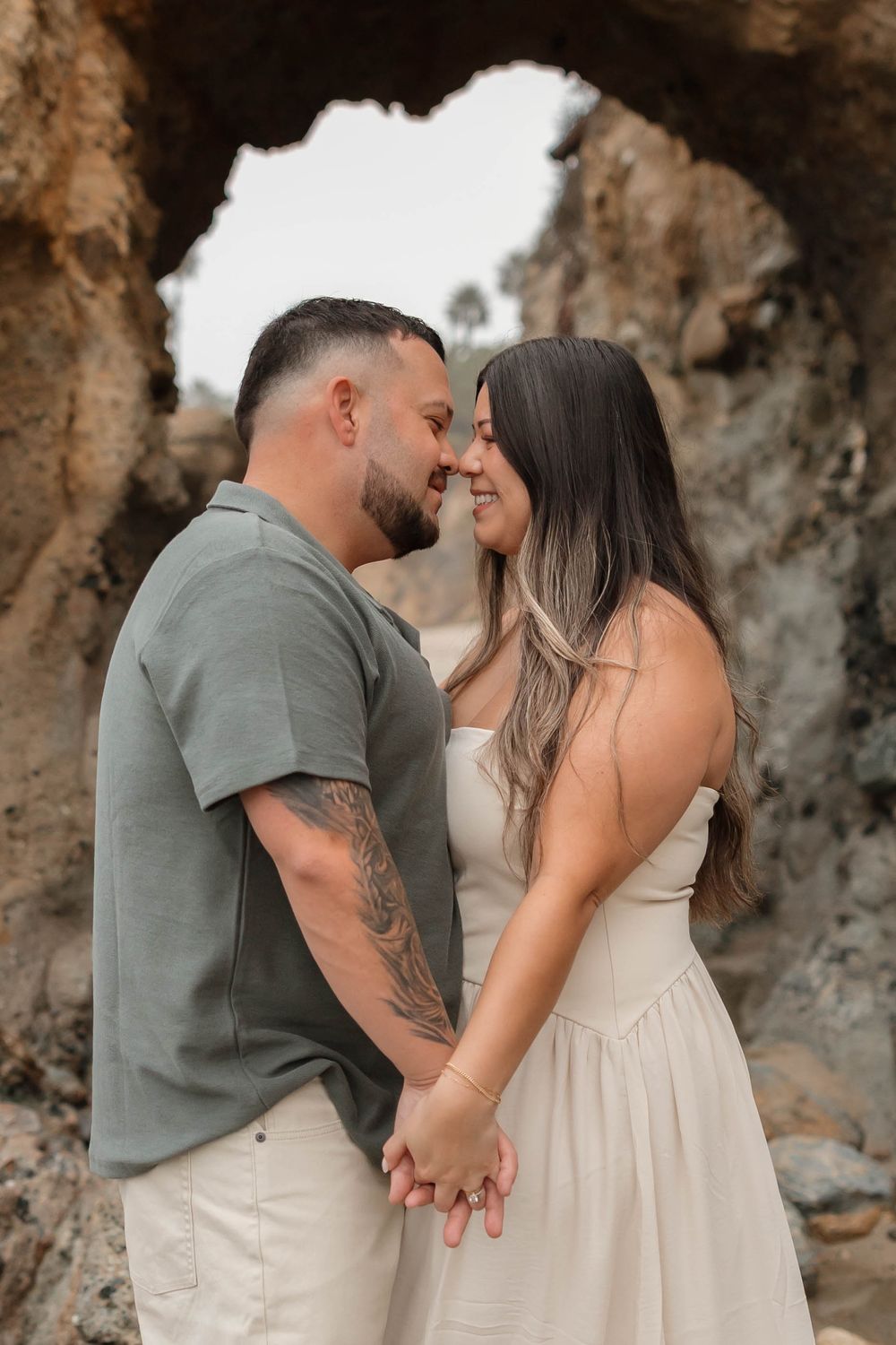 A romantic couple shares tender moments on a beach against a rocky cliff backdrop in matching cream and sage outfits.
