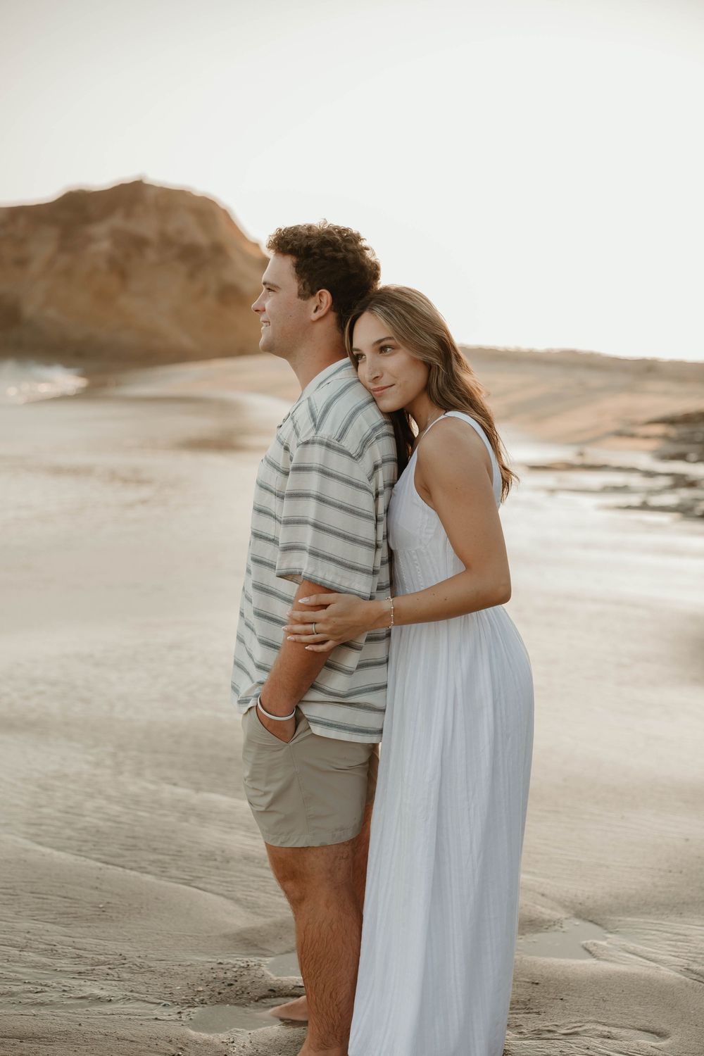 A couple embraces tenderly on a serene beach at sunset with cliffs in the background.