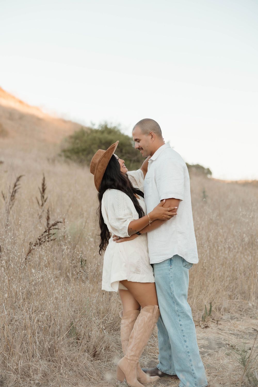 Couple embracing in golden field, woman in white dress and cowboy hat, man in white shirt and jeans, sunset hills backdrop.
