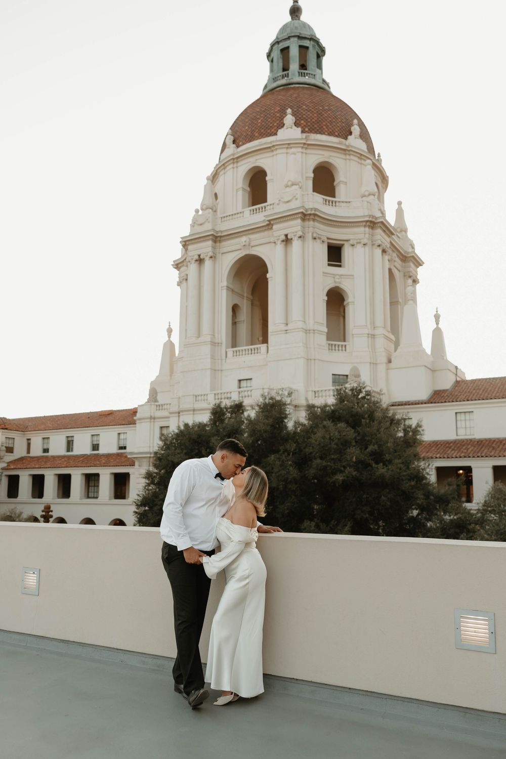 Couple embraces on rooftop terrace with beautiful Pasadena City Hall dome and architecture in background.