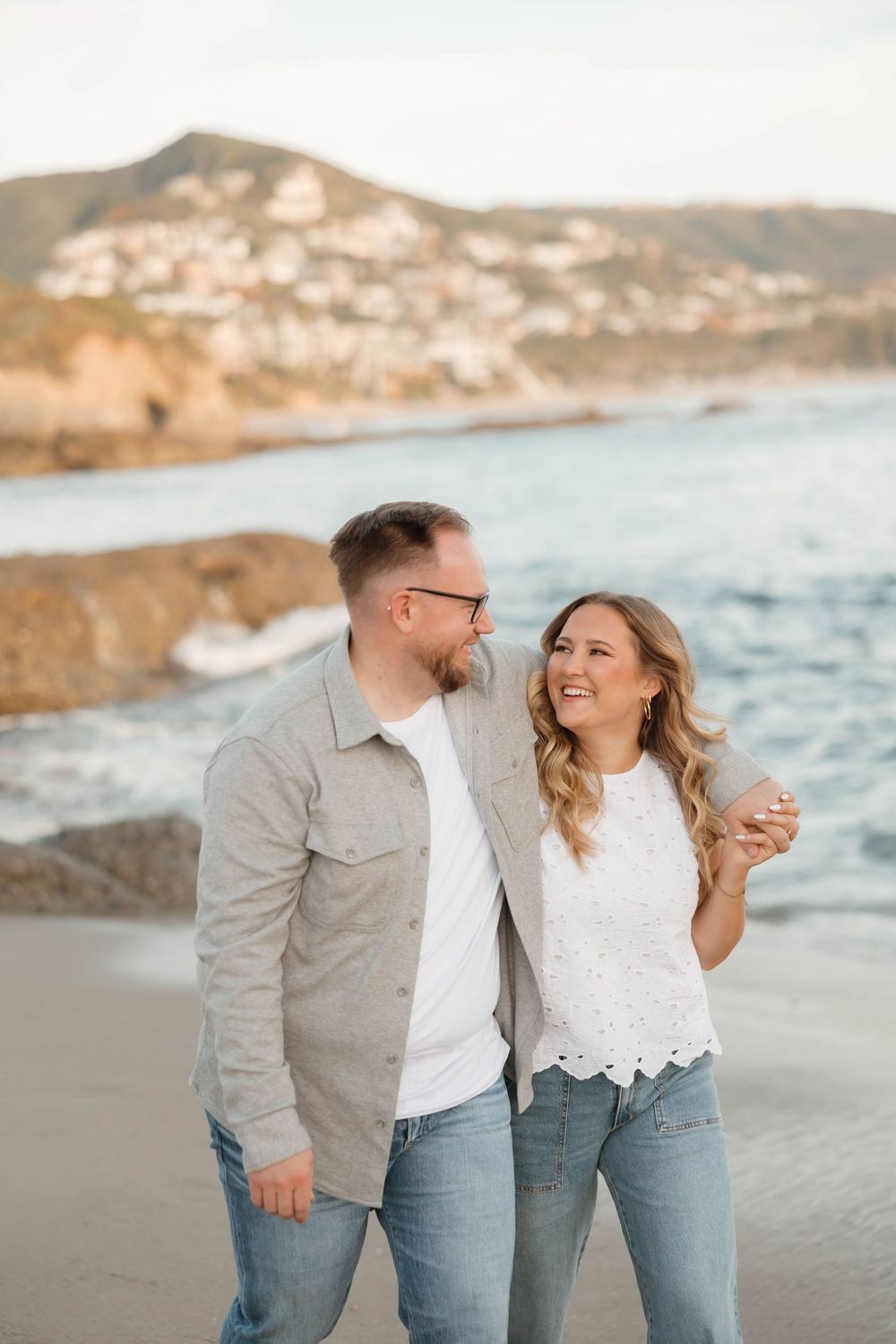 A smiling couple walks along a rocky beach with ocean waves and a hillside town in the background.