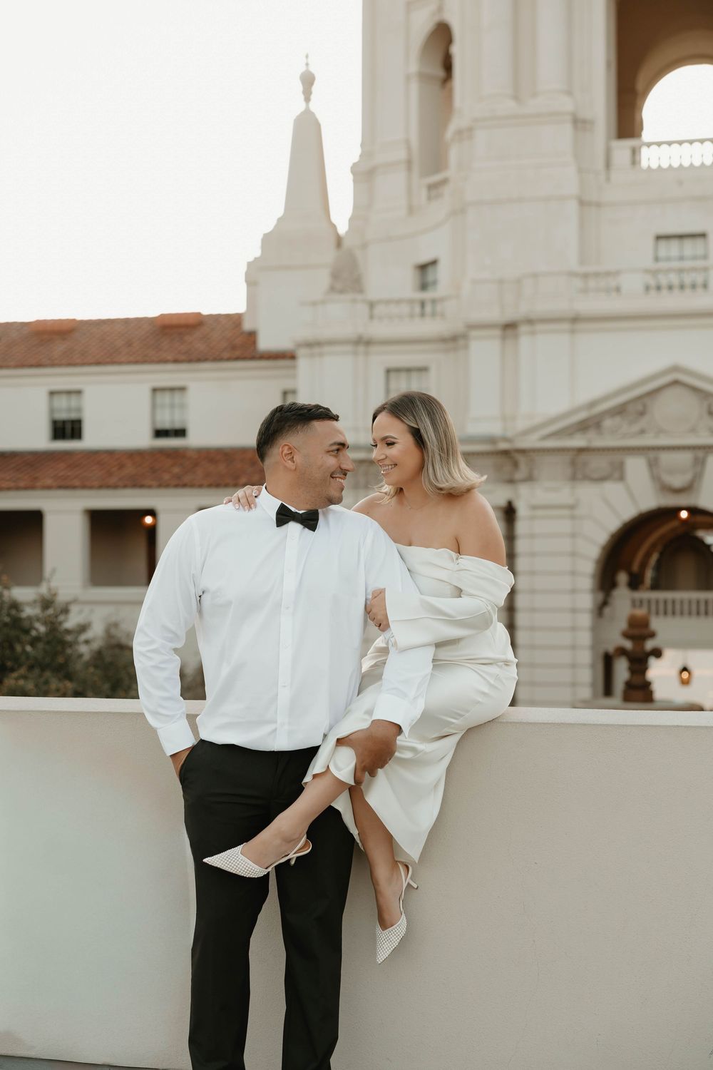 A romantic couple in formal attire share an intimate moment on a balcony of an elegant white building at sunset.