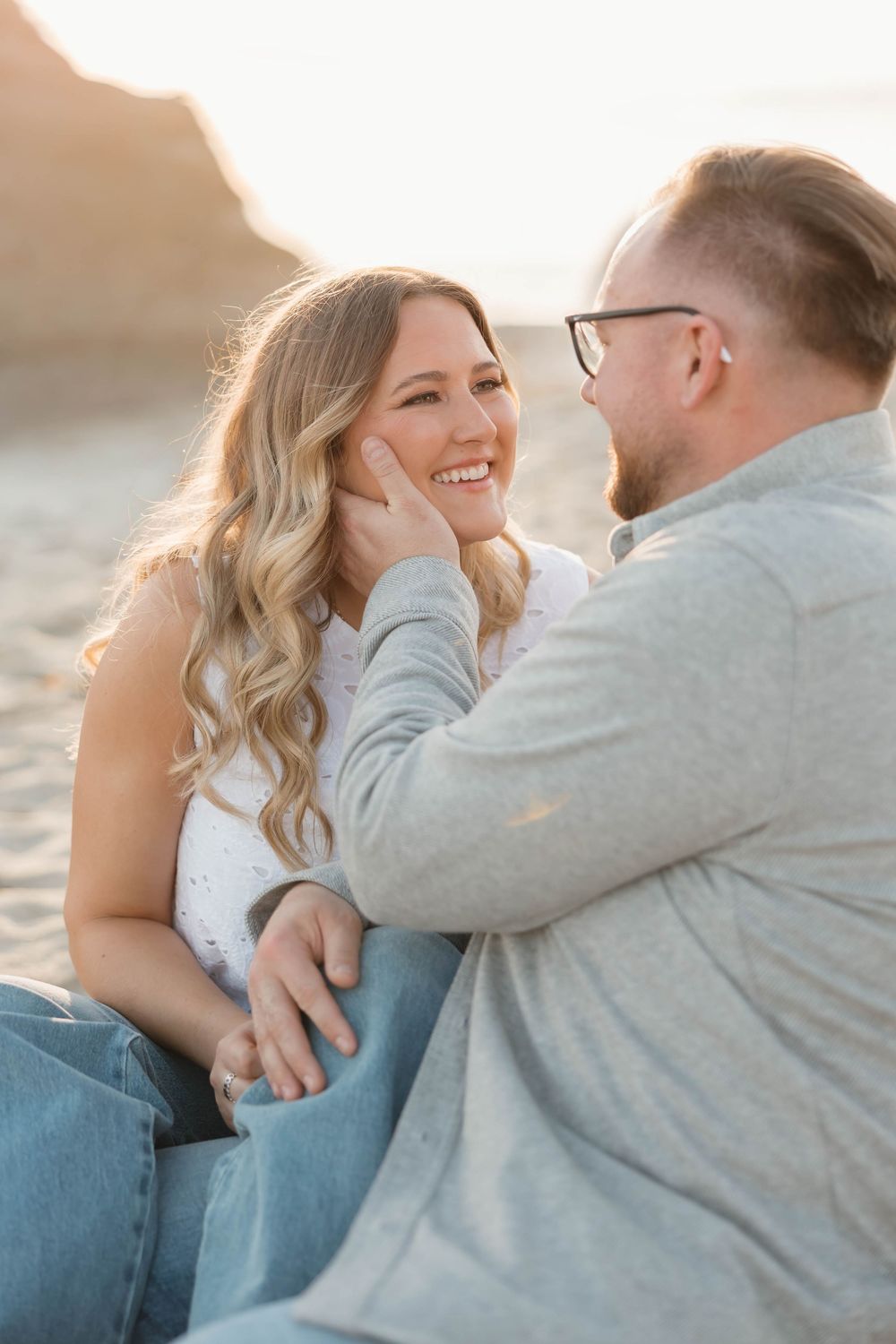 A couple shares a tender moment on the beach at golden hour, the man gently cupping the woman's smiling face.