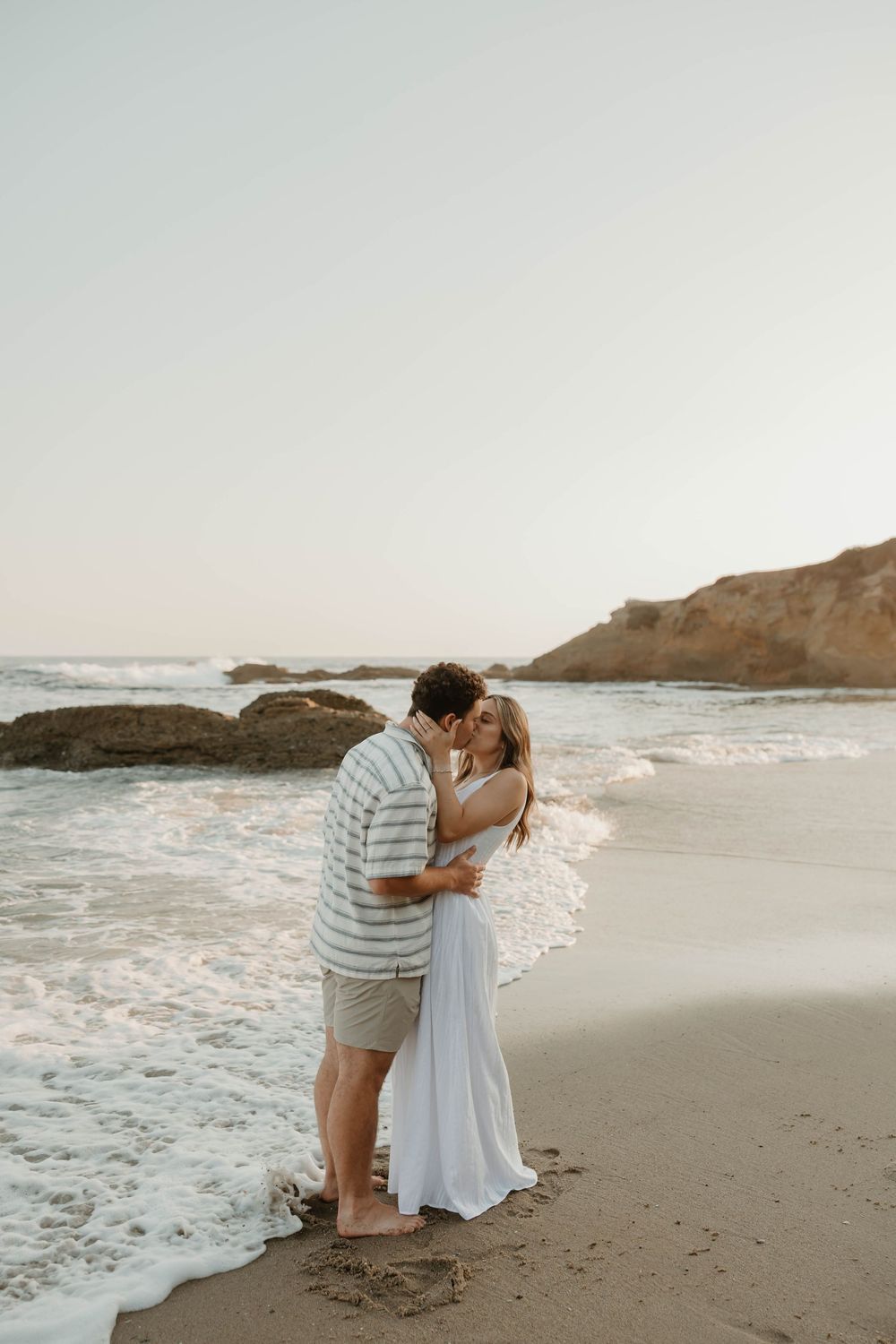 A romantic couple shares intimate moments on a serene beach at sunset with waves crashing in the background.
