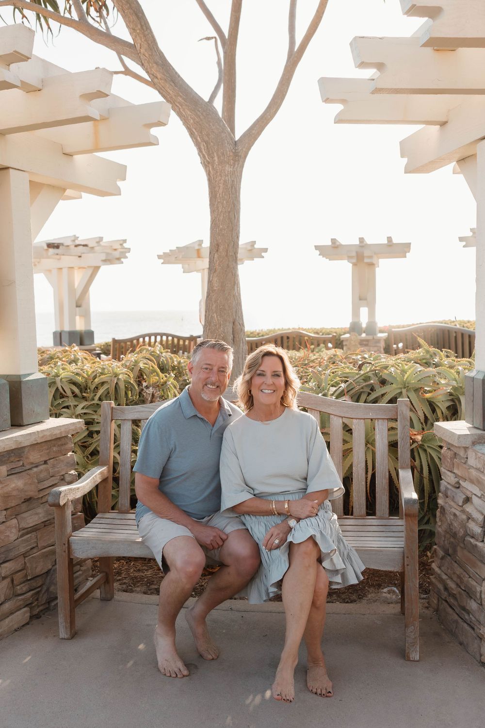 Smiling couple sitting on a wooden bench under a pergola with ocean views and lush greenery in the background.