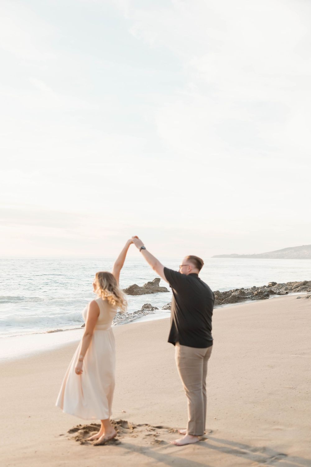 Couple dancing on a beach at sunset, man spinning woman in white dress near ocean waves.