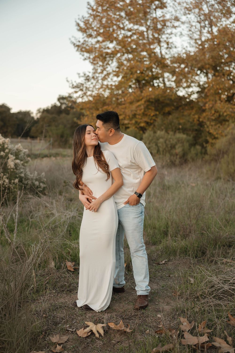 Romantic couple embraces in a golden autumn field during sunset while wearing white and light colored casual outfits.