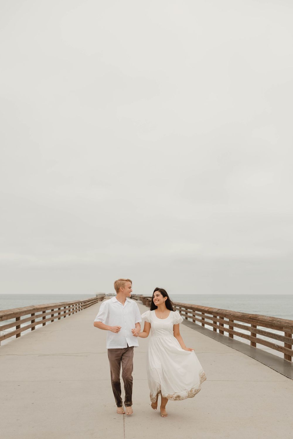 A couple in white attire walks together on a wooden pier during an overcast day at the beach.