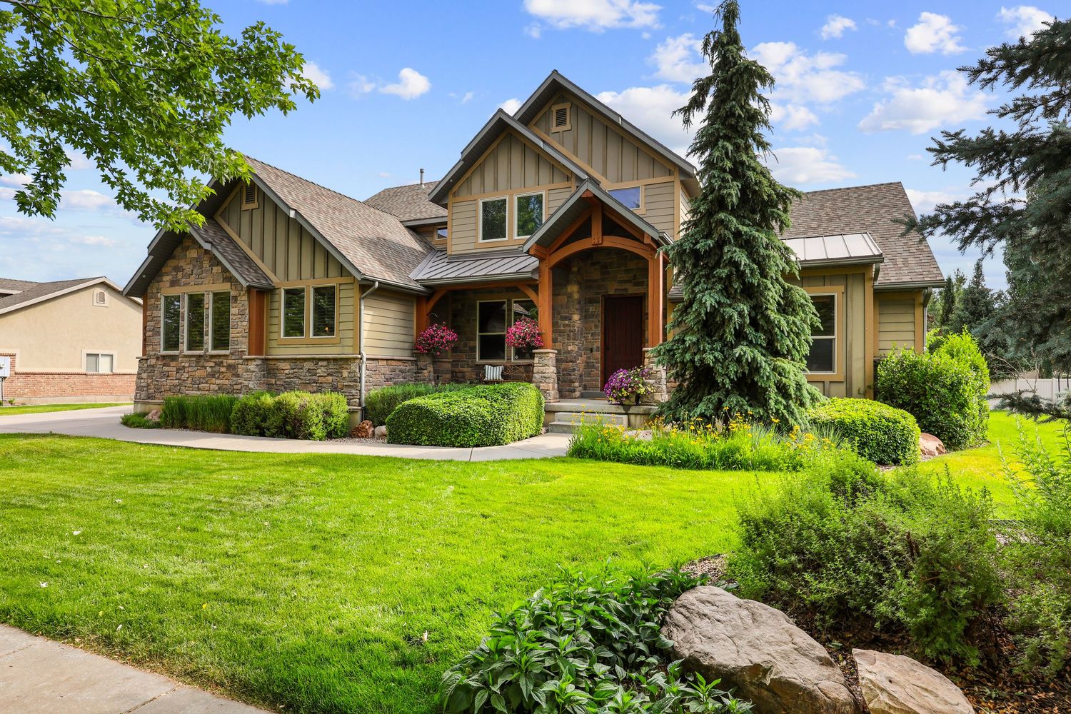 Craftsman-style home with stone accents, lush green lawn, mature trees, and inviting front entry. Captured by Slate Media (best in UT)