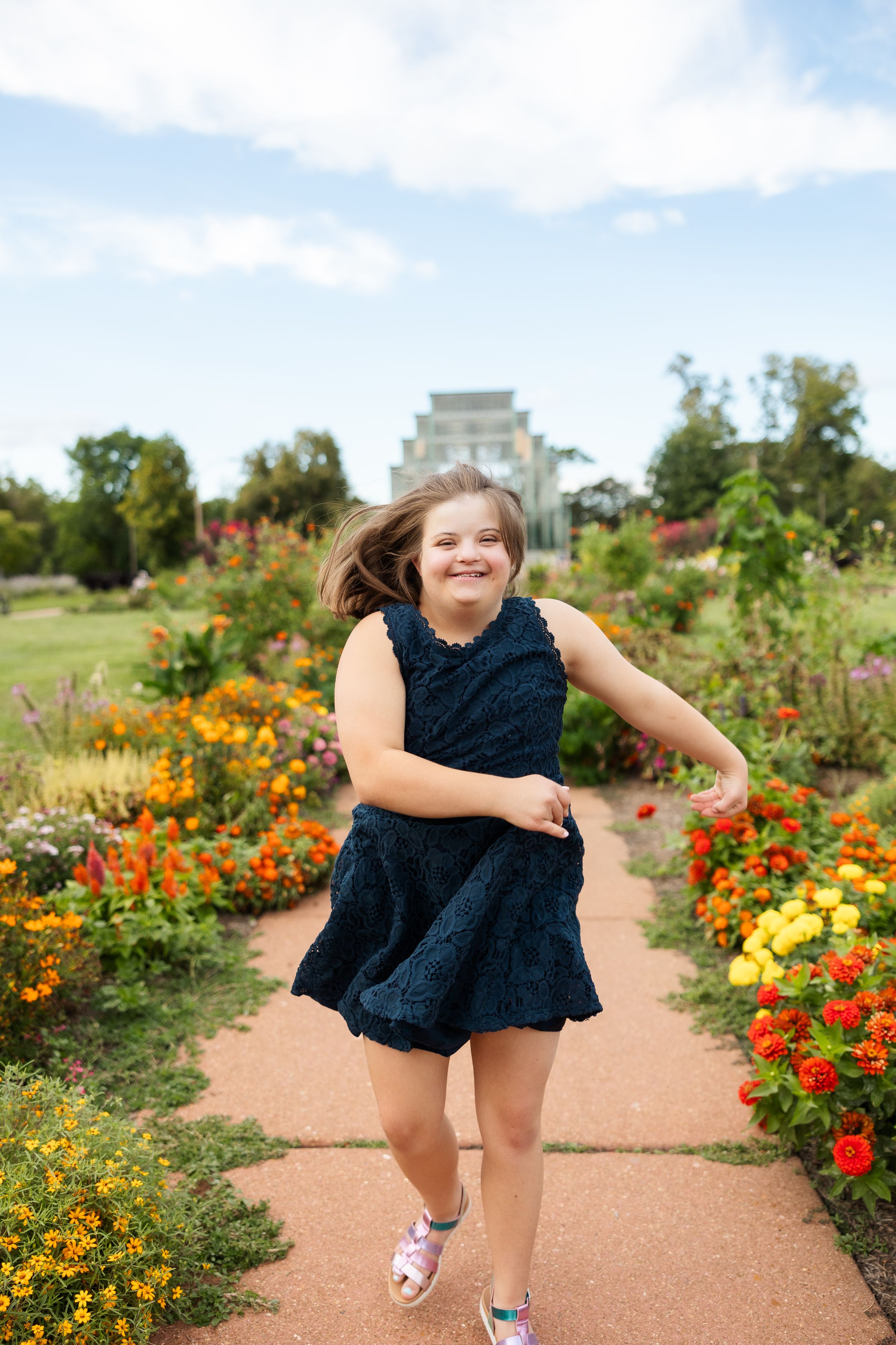 Girl in navy dress jumping joyfully on garden path surrounded by colorful flowers.