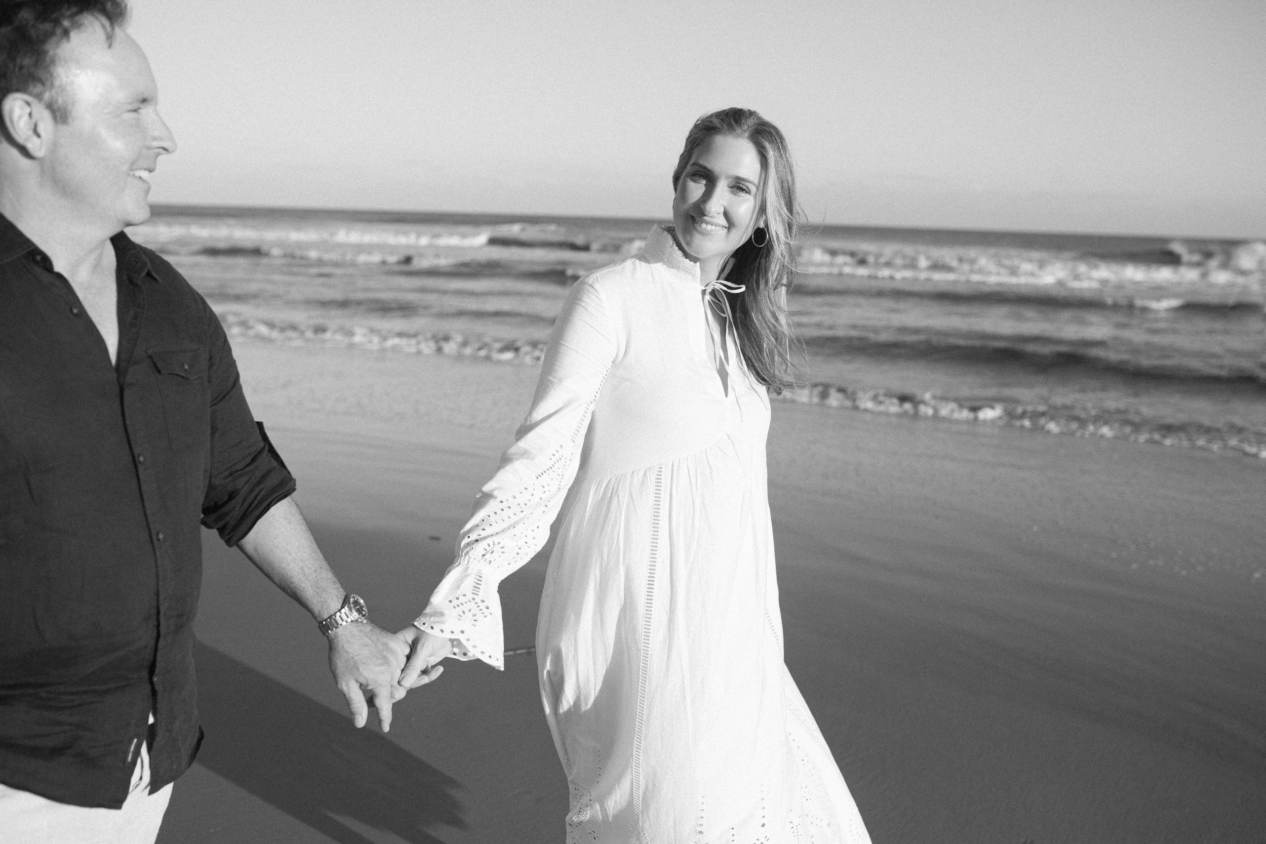 Black and white photo by B. Logsdon of a couple holding hands while walking on a beach with ocean waves of HHI, SC in background.