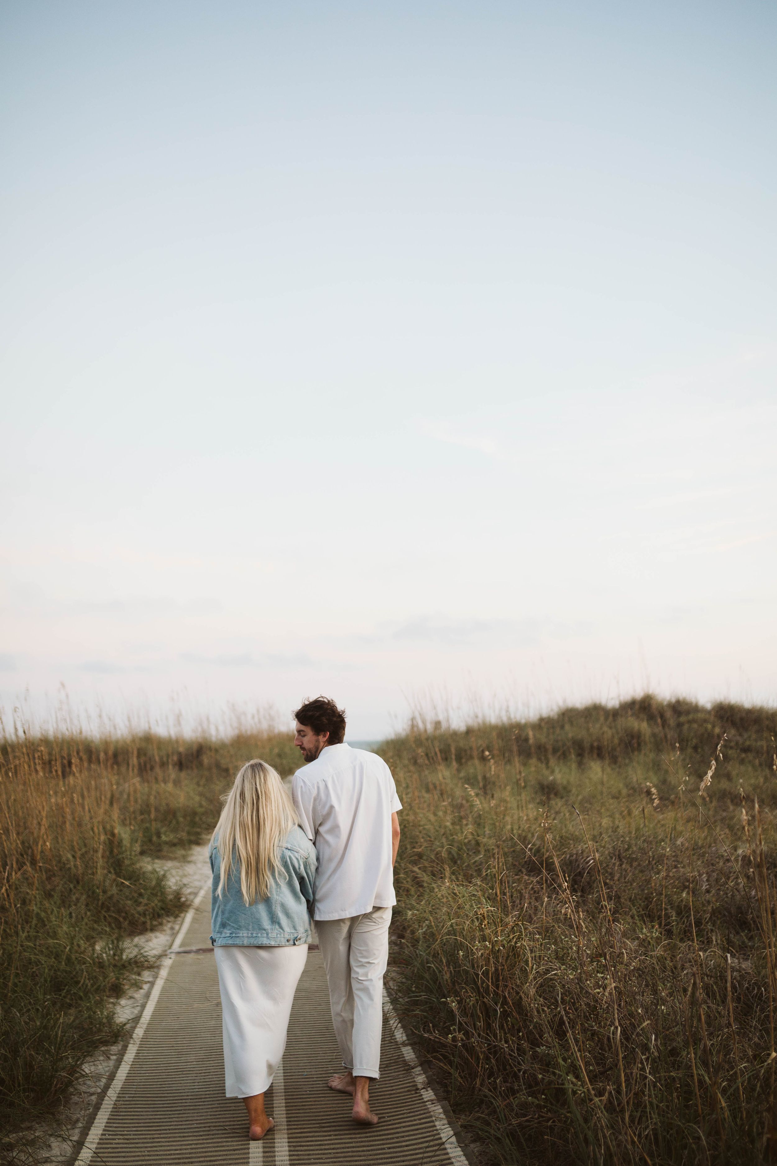 Photo by B.Logsdon of two people walking together on a boardwalk path through tall coastal grass at dusk on Hilton Head Island, SC.