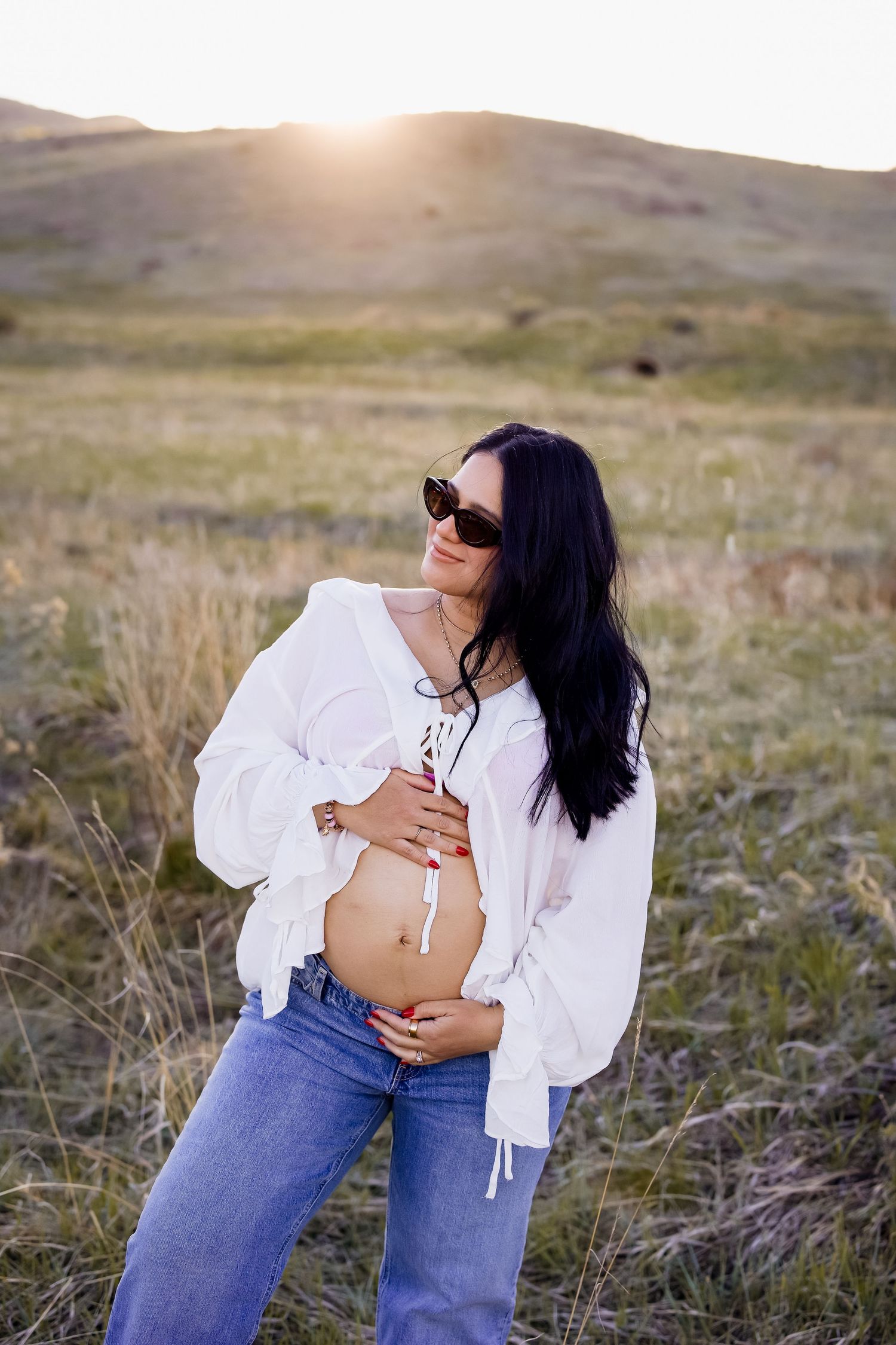 Pregnant woman in white blouse and jeans cradling her baby bump at a golden sunset outdoors.