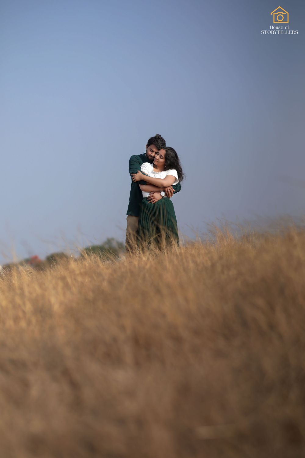 A romantic couple embraces in a golden grass field against a blue sky backdrop.
