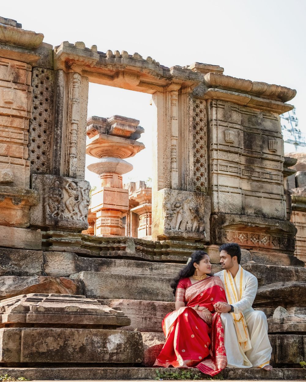 A couple in red and beige outfits share romantic moments against ancient stone ruins during a sunset photoshoot.
