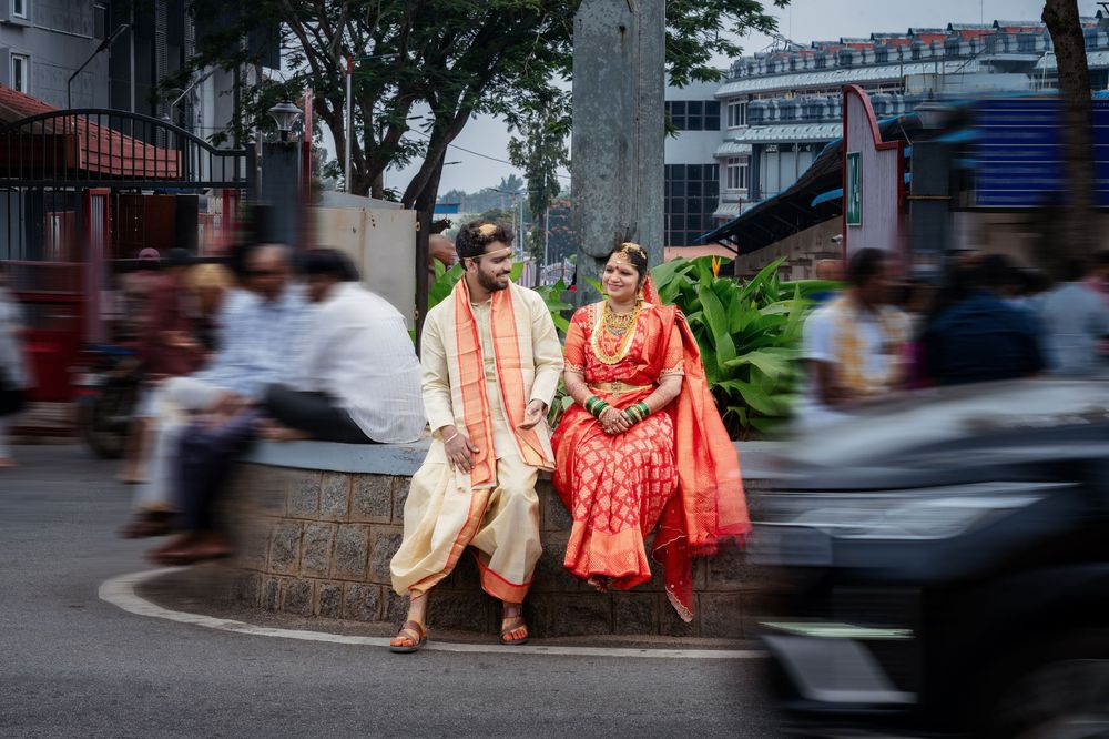 Indian couple in traditional wedding attire posing on a busy street, bride in red saree and groom in cream dhoti.