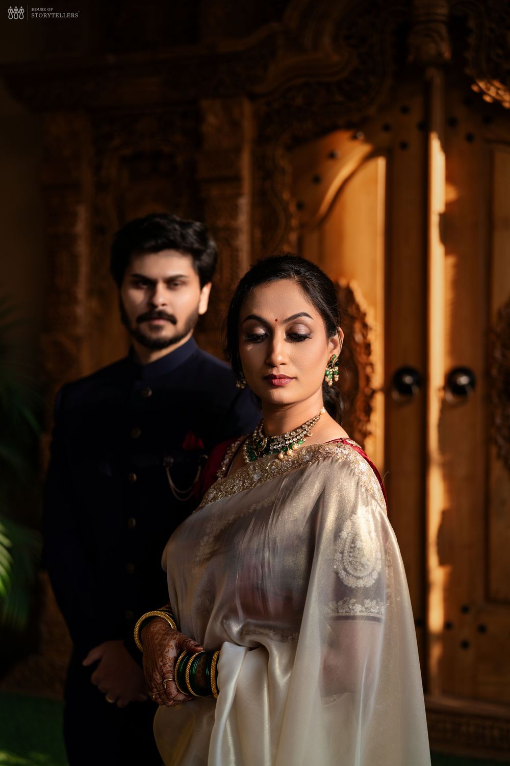 Bride in white embroidered saree with gold jewelry stands before groom in dark sherwani at traditional Indian wedding.