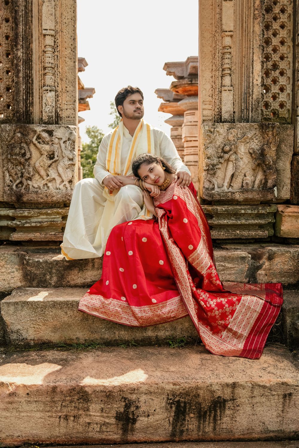 Couple in traditional Indian attire sitting on ancient temple steps, bride in red saree, groom in white kurta.