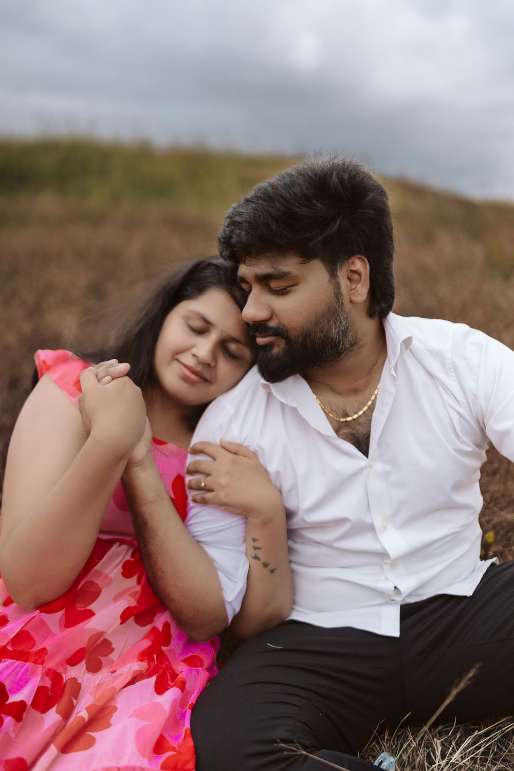 A couple sitting outdoors, woman in pink floral dress leaning on man in white shirt, sharing a tender romantic moment.