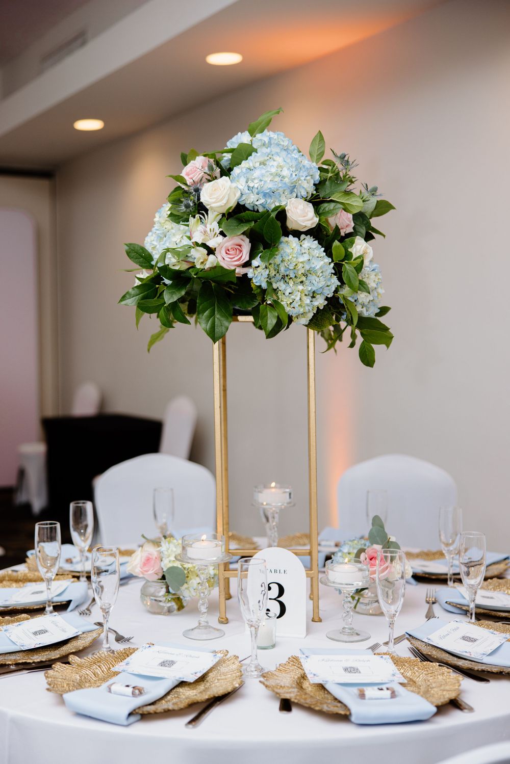 Elegant wedding table centerpiece with tall gold stand featuring blue hydrangeas and pink roses atop a white tablecloth.