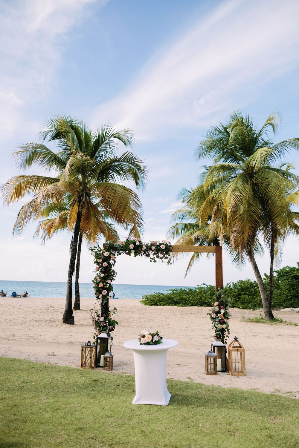 Romantic beach wedding arch with palm trees and lanterns on a tropical beach setup with white decor.