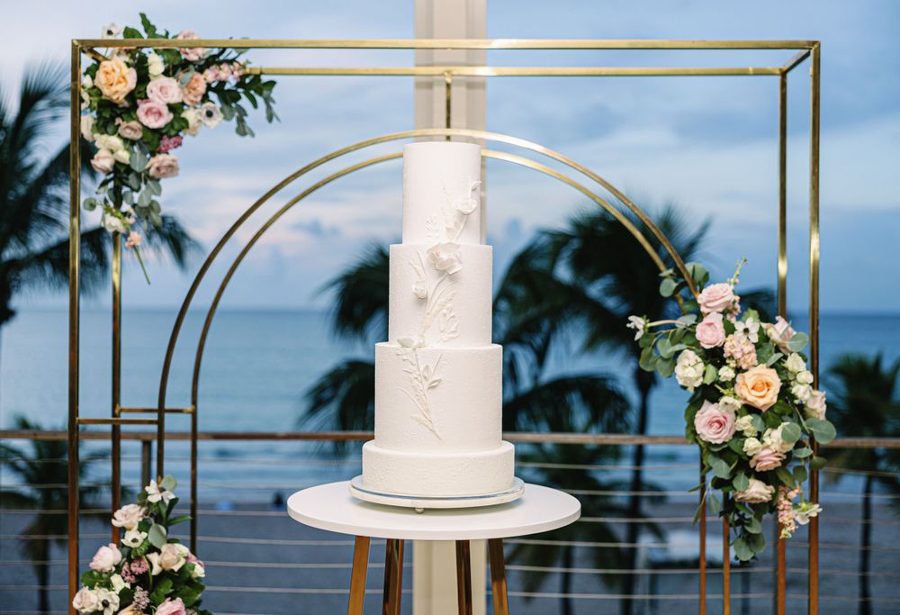 Elegant tiered white wedding cake displayed on modern geometric arch with floral decorations.