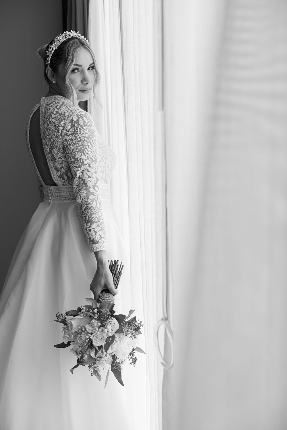 Elegant bridal portrait by window light showcasing lace gown and bouquet.