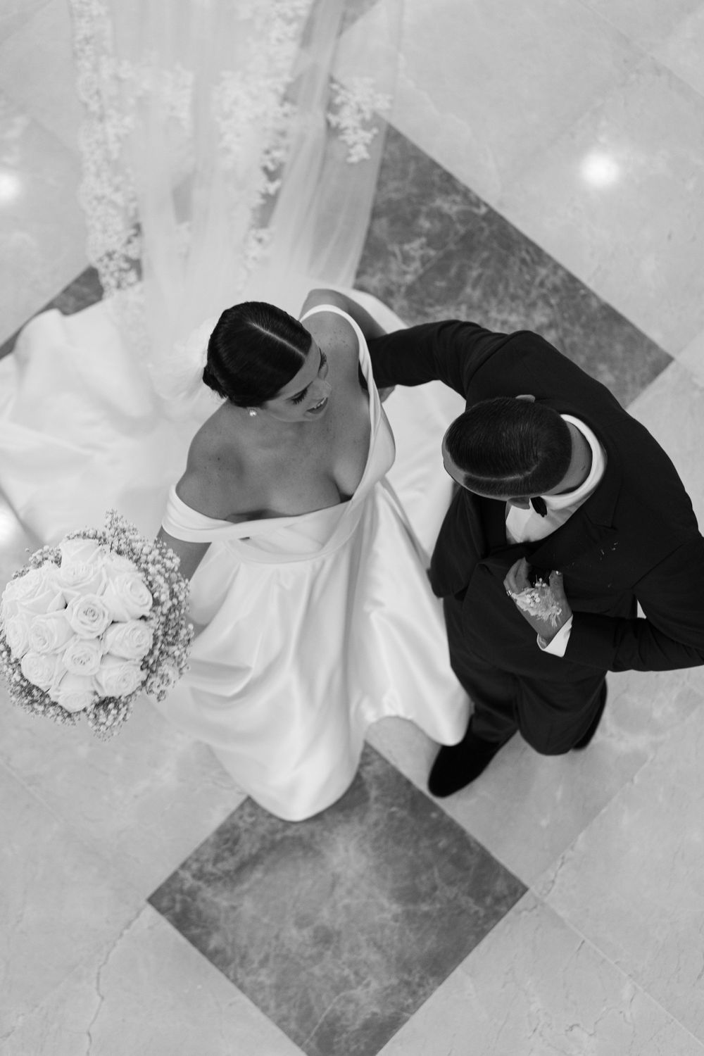 An overhead black and white view of a couple in formal attire with a white bouquet on a tiled floor.