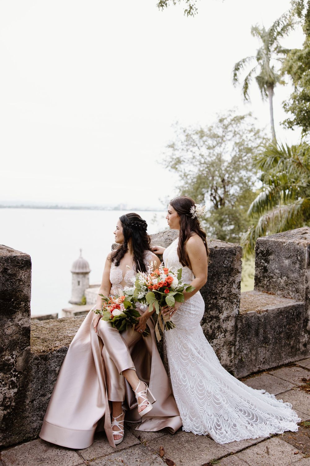 Two people in elegant dresses pose with colorful bouquets against a historic stone wall by the ocean.