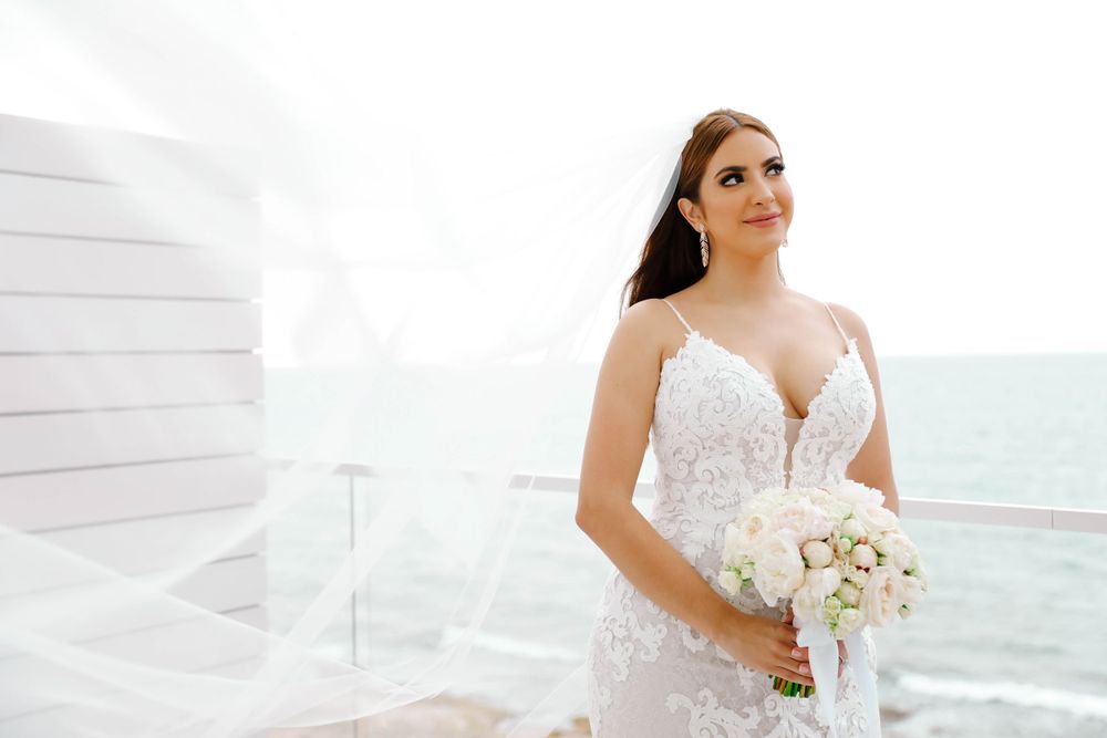 Bride in lace wedding dress holds white bouquet while standing on oceanfront balcony.
