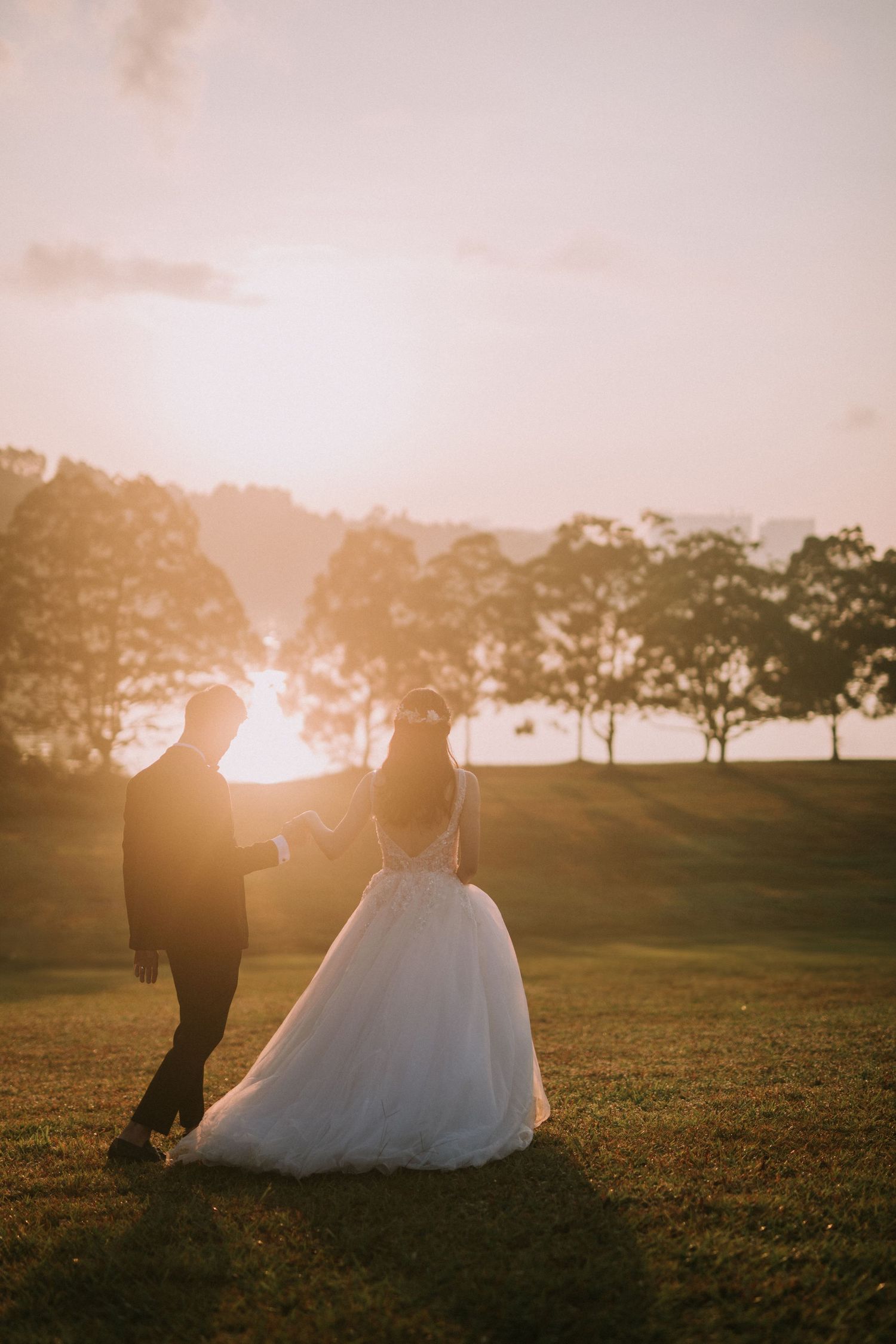 Newlyweds walk through a sunlit field at golden hour creating beautiful silhouettes.