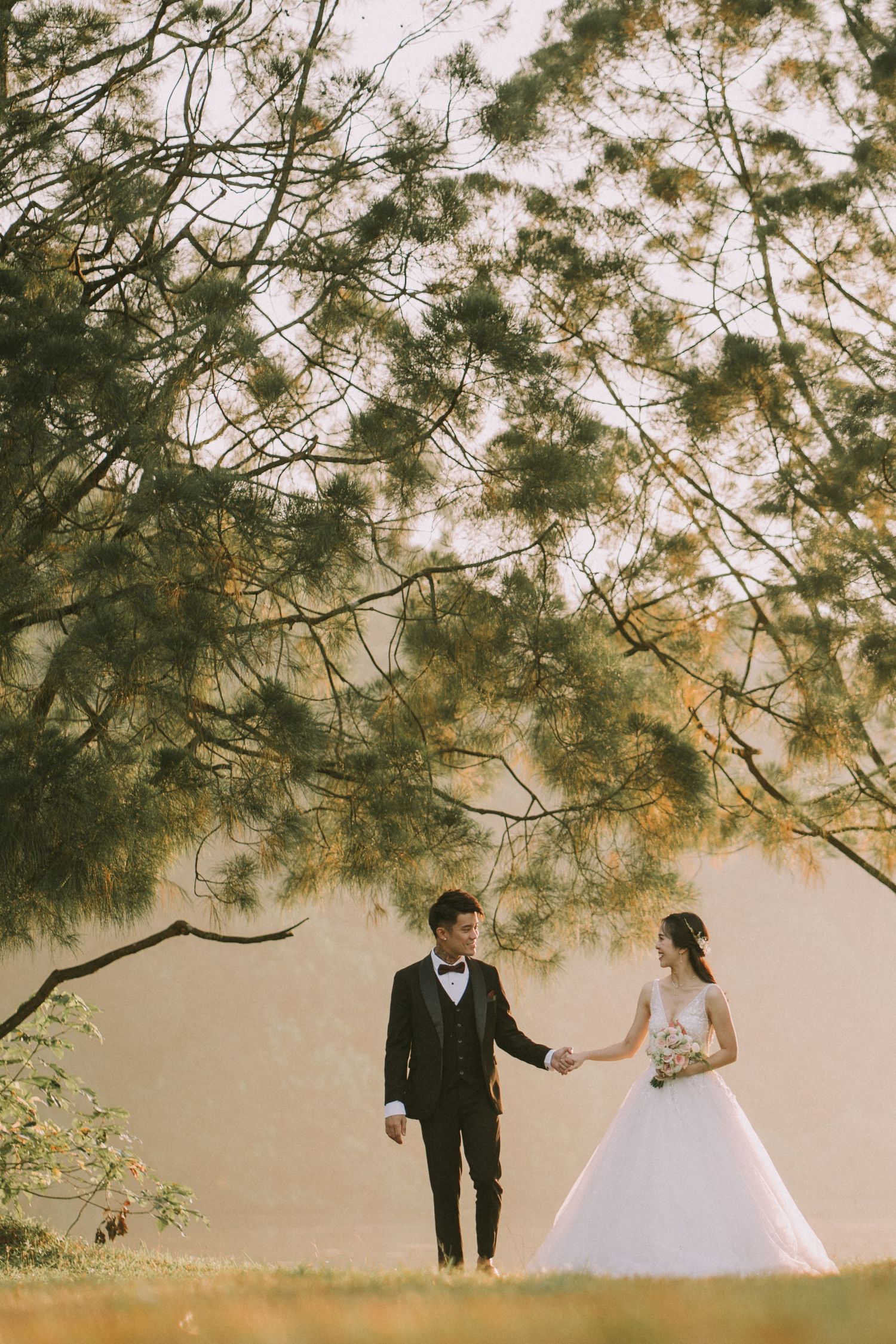 A wedding couple shares a romantic moment under overhanging tree branches by the water.