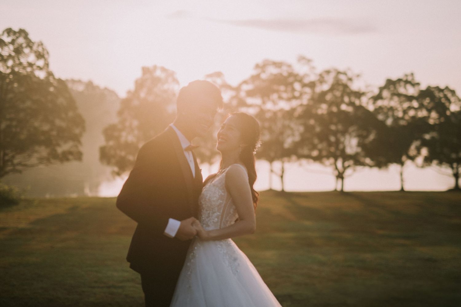 Newlyweds embrace in golden hour lighting with trees in the background.