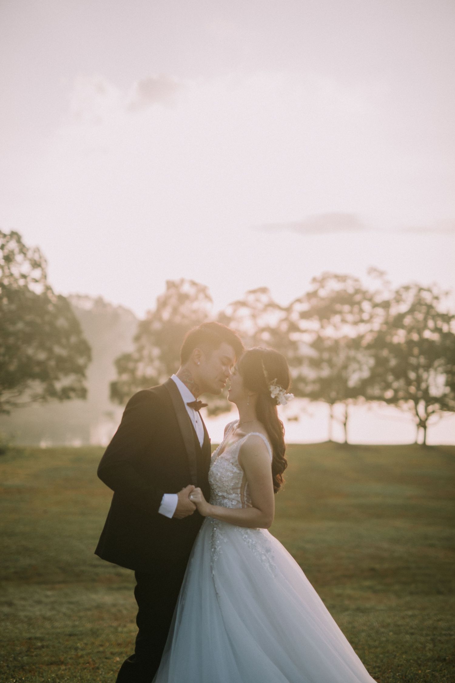 A couple in wedding attire poses together on a grassy hill at sunset.