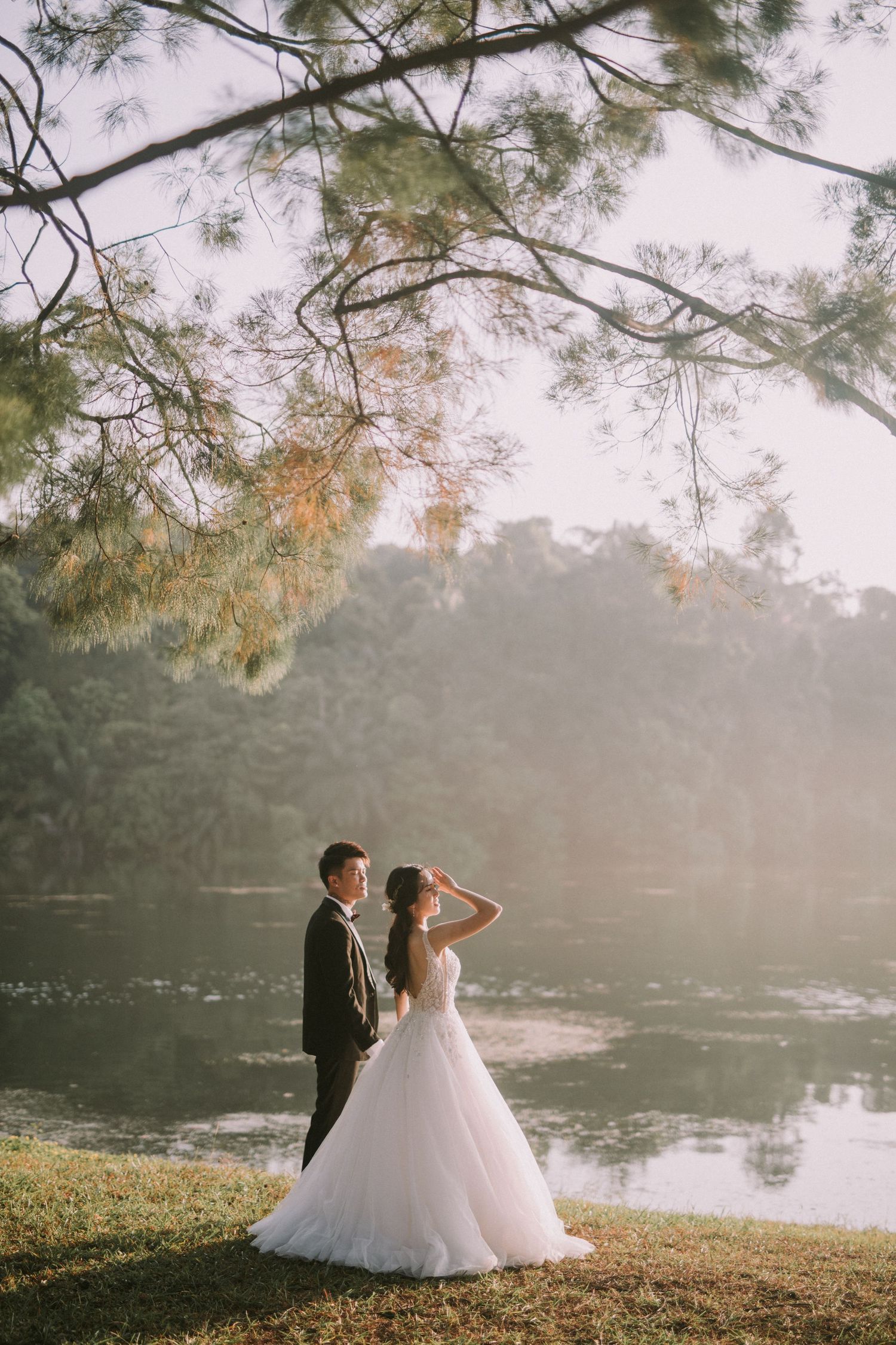 A wedding couple stands by a serene lake with overhanging tree branches.