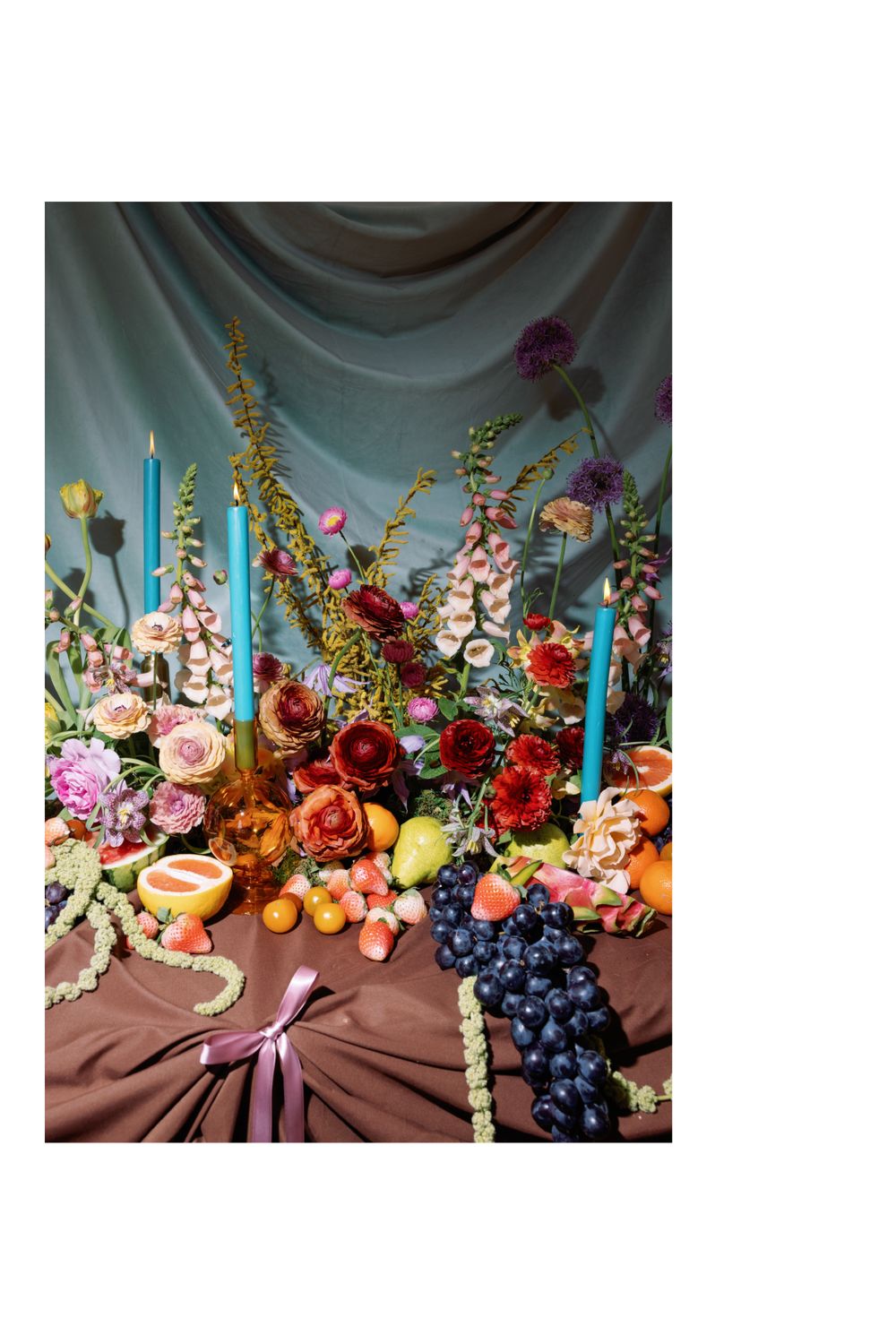 Dramatic floral still life with foxgloves, roses, and fresh fruit arranged against a blue draped backdrop.