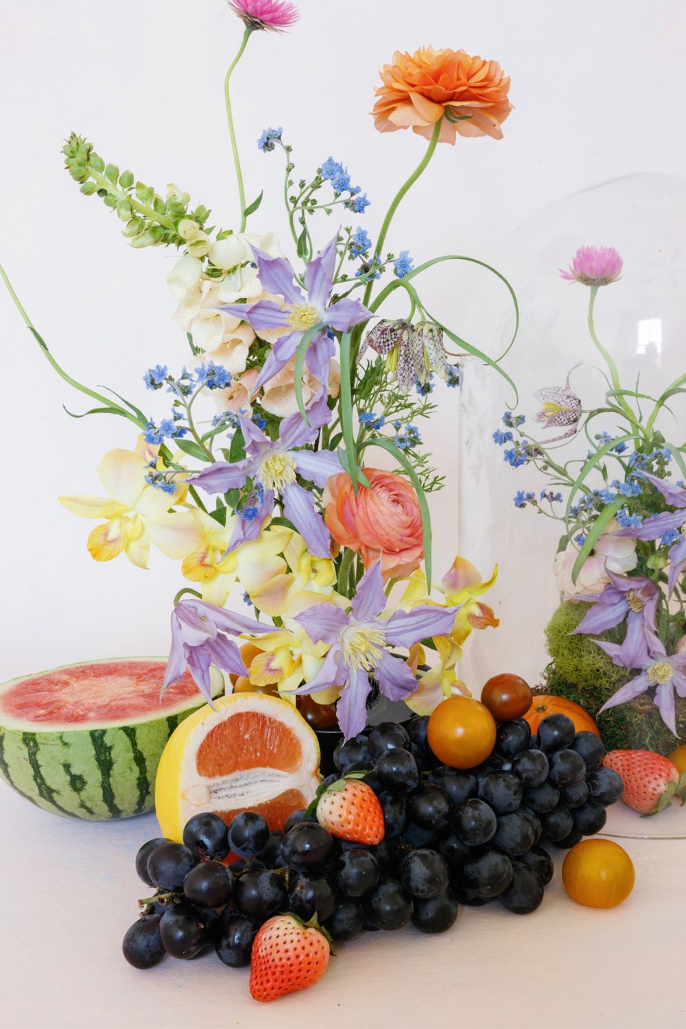 Fresh flower arrangement with purple clematis and citrus fruits alongside watermelon and grapes on white surface.