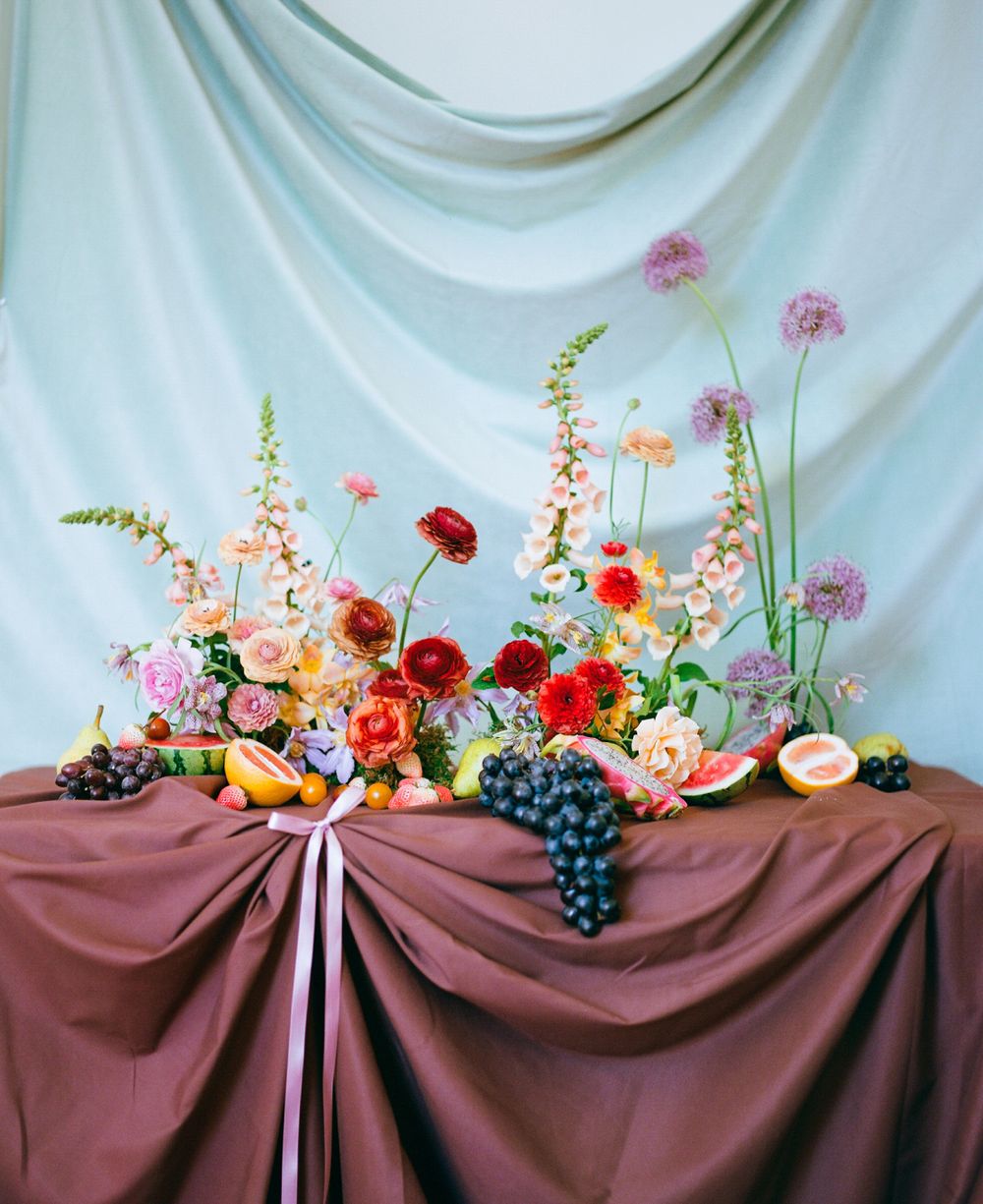 Elegant floral centerpiece with roses, ranunculus, and fruits against a burgundy draped table.