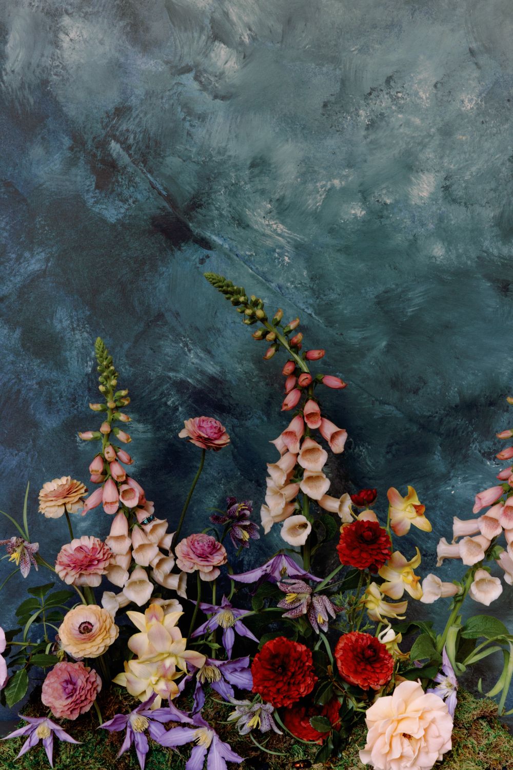 Romantic floral arrangement with foxgloves and roses against dramatic blue-green textured background.