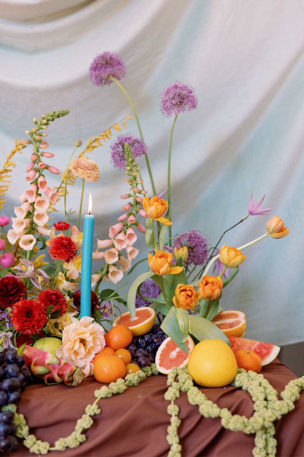 Elegant floral centerpiece with pink foxgloves, citrus fruits, and purple alliums on draped fabric.