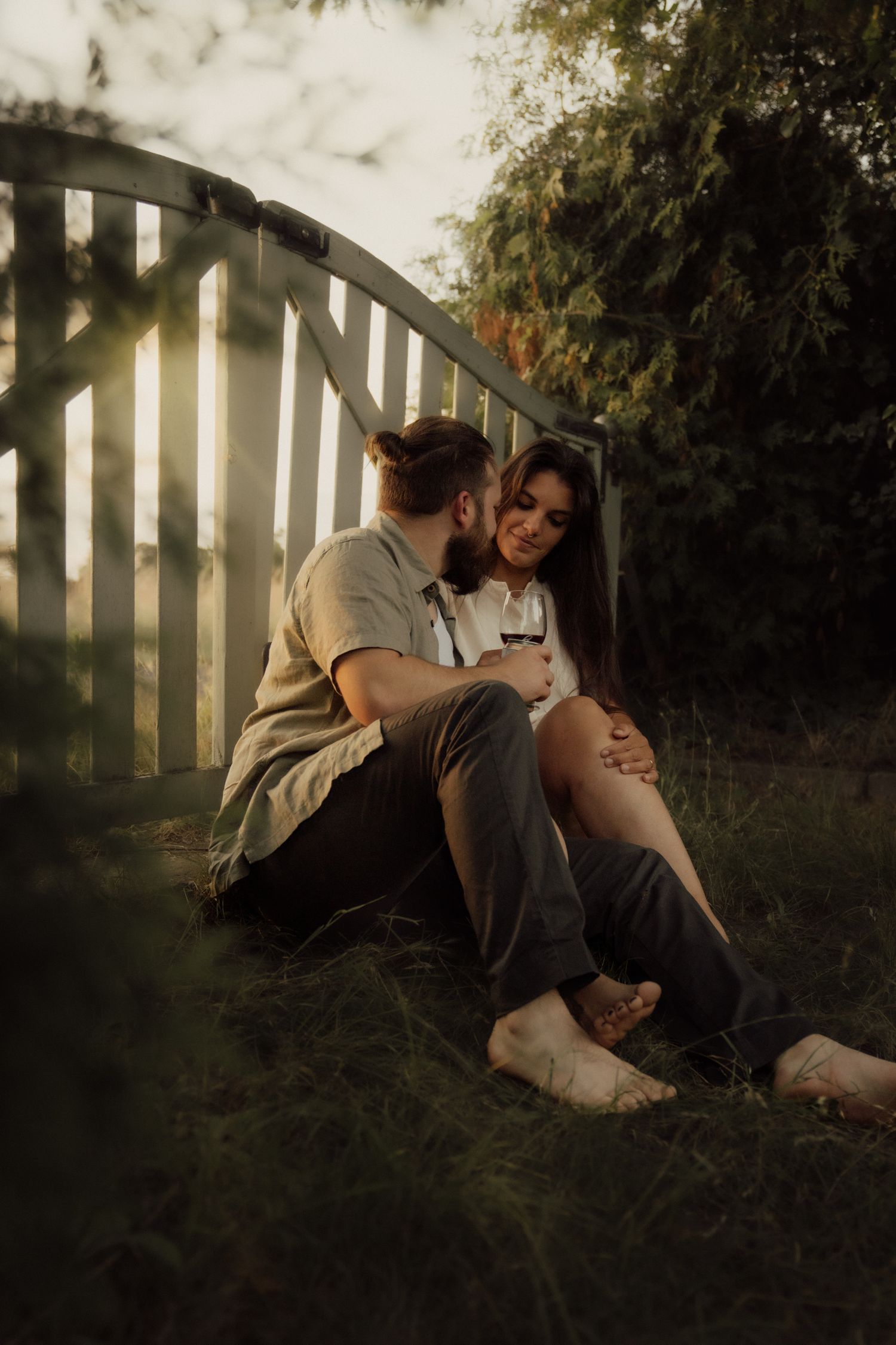 Couple sitting together on grass near a curved wooden fence at golden hour.