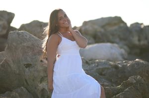 Woman in flowing white dress poses elegantly among coastal rocks at sunset.