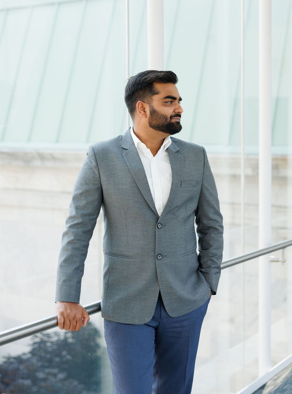 Bearded man in gray blazer and blue trousers stands by glass railing in modern building, looking away.