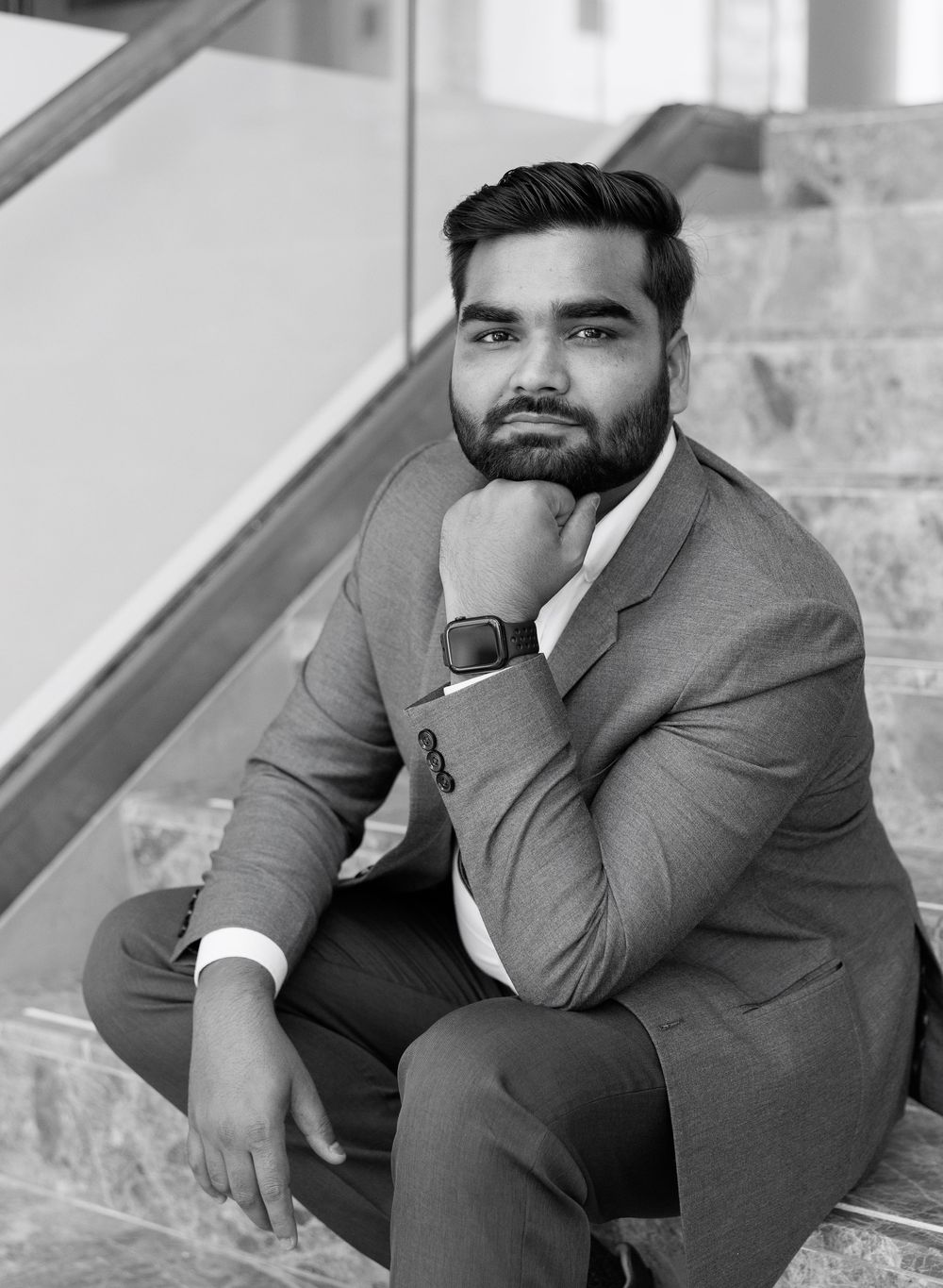 A well-dressed bearded man in a gray suit sits thoughtfully on stairs in a black and white professional portrait photo.